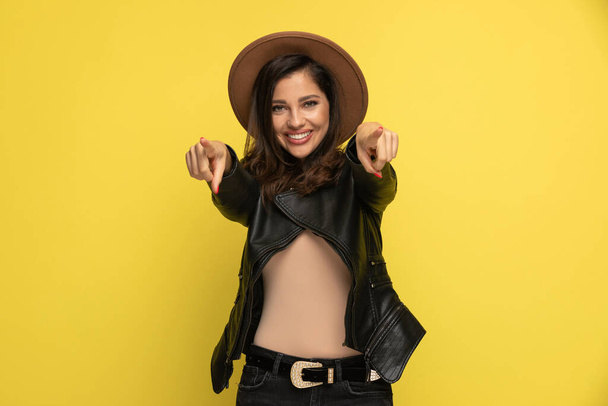 happy young girl in leather jacket pointing fingers and smiling on yellow background - Фото, изображение