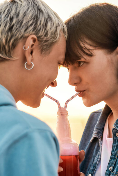 Close up of young lesbian couple looking at each other, drinking from one glass bottle with the straw, Two women enjoying cold beverage on a summer day outdoors - 写真・画像