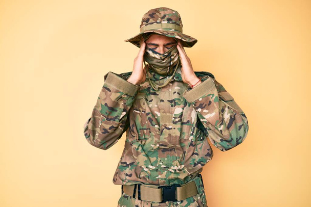Fiatal jóképű férfi, aki álcázott katonai egyenruhát és maszkot visel, fejfájásban szenved, kétségbeesetten és stresszesen, mert fáj és migrén van. kezeket a fejre..  - Fotó, kép