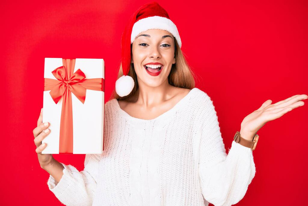Genç esmer kadın Noel şapkası takıyor ve elinde zaferi kutlayan bir hediye var. Mutlu bir gülümseme ve ellerini kaldırarak kazanan bir ifade.  - Fotoğraf, Görsel