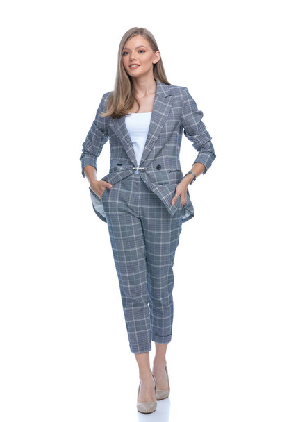 νεαρή επιχειρηματίας σε μπλε καρό κοστούμι κρατώντας τα χέρια στις τσέπες, χαμογελώντας και περπατώντας απομονωμένη σε λευκό φόντο, πλήρες σώμα - Φωτογραφία, εικόνα
