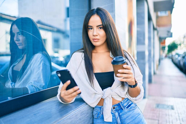 Jeune fille hispanique avec une expression sérieuse en utilisant un smartphone et boire du café à emporter à la ville. - Photo, image