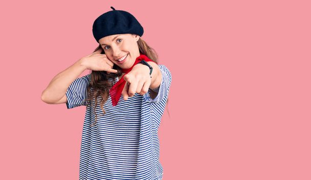 Young beautiful blonde woman wearing french beret and striped t-shirt smiling doing talking on the telephone gesture and pointing to you. call me.  - Photo, Image