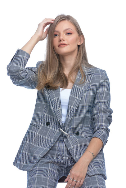 Ευτυχισμένη νεαρή επιχειρηματίας σε μπλε καρό κοστούμι τακτοποίηση των μαλλιών και χαμογελώντας, κάθεται απομονωμένη σε λευκό φόντο - Φωτογραφία, εικόνα