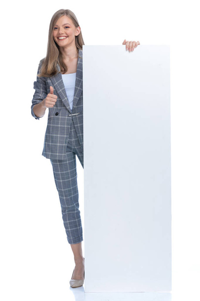 Ευτυχισμένη νεαρή επιχειρηματίας σε μπλε καρό κοστούμι κάνοντας τους αντίχειρες επάνω χειρονομία και παρουσιάζοντας άδειο πίνακα, χαμογελώντας και στέκεται απομονωμένη σε λευκό φόντο, πλήρες σώμα - Φωτογραφία, εικόνα
