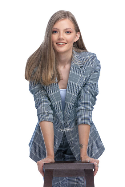 happy young girl in blue checkered suit leaning on wooden chair and smiling, standing isolated on white background - Фото, изображение