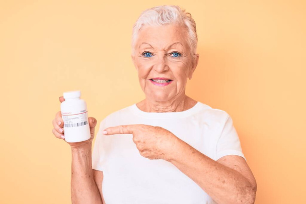 Senior beautiful woman with blue eyes and grey hair holding pills smiling happy pointing with hand and finger  - Zdjęcie, obraz
