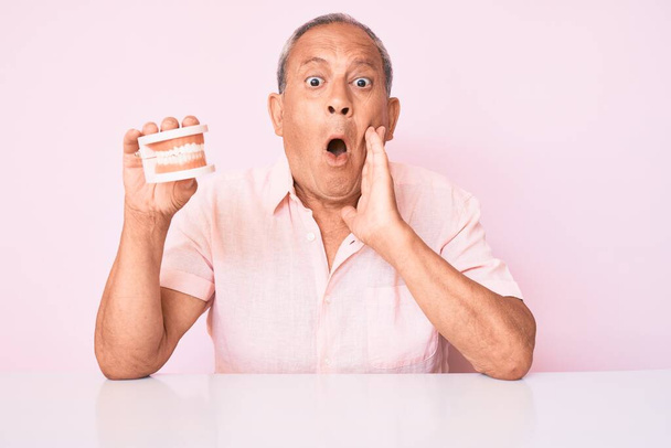 Senior handsome man with gray hair holding denture sitting on the talbe scared and amazed with open mouth for surprise, disbelief face  - Photo, Image