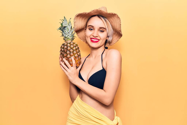 Jovem mulher loira bonita usando biquíni e chapéu de verão segurando abacaxi olhando positivo e feliz de pé e sorrindo com um sorriso confiante mostrando dentes  - Foto, Imagem