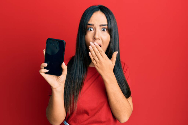 Νεαρή όμορφη ισπανόφωνη κοπέλα κρατώντας smartphone δείχνει οθόνη που καλύπτει το στόμα με το χέρι, σοκαρισμένος και φοβισμένος για λάθος. έκπληκτη έκφραση  - Φωτογραφία, εικόνα