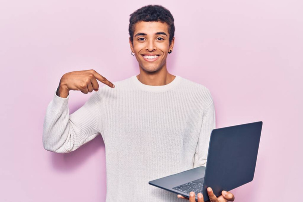 Молодой африканский американец держит ноутбук, указывая пальцем на себя, улыбающийся счастливый и гордый  - Фото, изображение