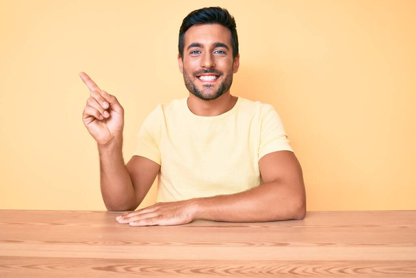 Fiatal spanyol férfi alkalmi ruhát visel ül az asztalon mosolyogva boldog rámutatva a kezét és ujját az oldalán  - Fotó, kép