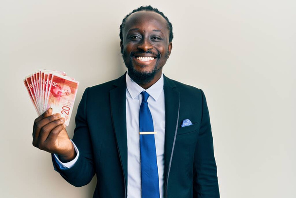 Guapo joven negro con traje de negocios que sostiene billetes de 20 shekels que se ven positivos y felices de pie y sonriendo con una sonrisa confiada que muestra los dientes  - Foto, imagen