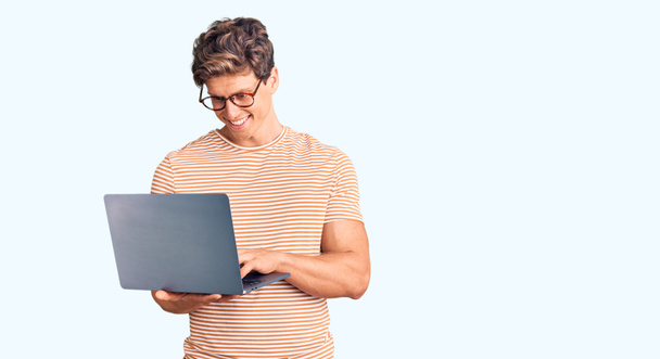 Joven hombre guapo con gafas que sostiene el ordenador portátil con aspecto positivo y feliz de pie y sonriendo con una sonrisa confiada que muestra los dientes  - Foto, imagen