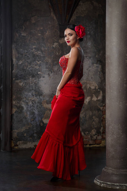 Portrait of a sultry brunette woman in a long red dress with a deep neckline and a red flower in her hair in the castle interior. - Photo, image