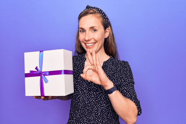 Young beautiful blonde woman holding birthday present box doing ok sign with fingers, smiling friendly gesturing excellent symbol  - Photo, image