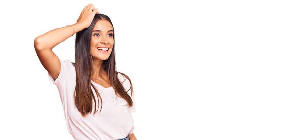 Молода іспаномовна жінка в повсякденній білій сорочці посміхається впевнено торкаючись волосся ручним жестом, позуючи привабливим і модним
  - Фото, зображення