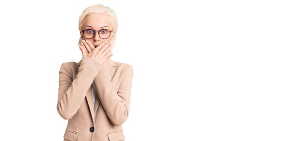 Young blonde woman wearing business clothes and glasses shocked covering mouth with hands for mistake. secret concept.  - Photo, image