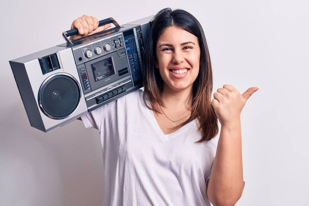 Young beautiful woman listening to music using vintage boombox over white background pointing thumb up to the side smiling happy with open mouth - Photo, Image