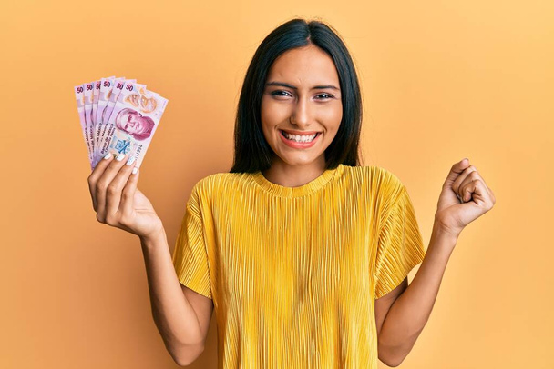 Junge brünette Frau mit 50 mexikanischen Pesos Banknoten schreit stolz und feiert Sieg und Erfolg sehr aufgeregt mit erhobenem Arm  - Foto, Bild