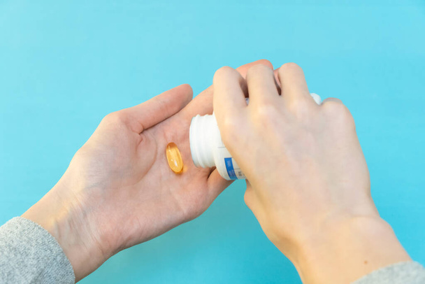 Una mano vierte cápsulas de vitaminas amarillas en la otra mano sobre un fondo azul - Foto, Imagen