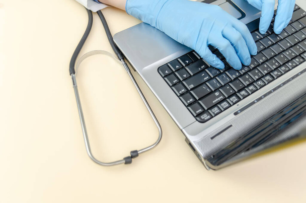 handen van een vrouwelijke arts in blauwe medische handschoenen werken op een laptop close-up, naast een stethoscoop. - Foto, afbeelding
