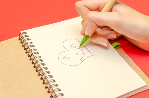 Main d'une fille qui dessine un cœur dans un carnet sur fond rouge. Concept de Saint Valentin, vacances - Photo, image
