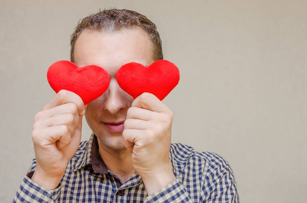 Elinde iki küçük kırmızı kalple gülümseyen koyu renk saçlı çekici bir adam gözlerini kağıt kalp sembolleriyle kaplıyor, açık bir arkaplanda duruyor. Sevgililer Günü - Fotoğraf, Görsel