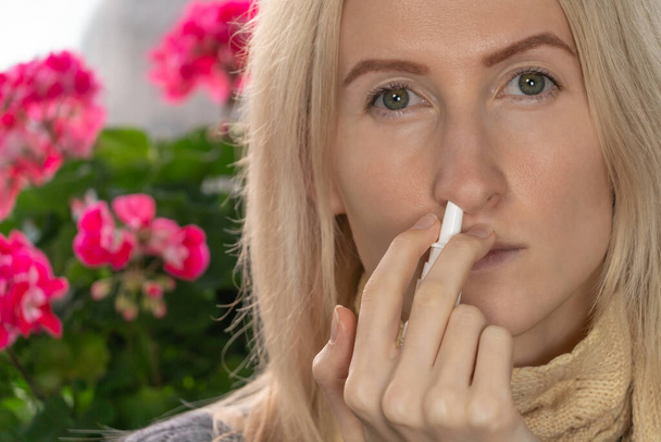 giovane donna bionda utilizza uno spray nasale per un naso che cola o allergia su uno sfondo di fiori al coperto, copiare lo spazio. - Foto, immagini