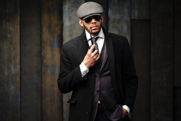 Studio shot of stylish african american man wearing coat, cap, glasses and holding smoking pipe - Foto, imagen