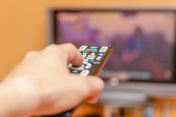 Το χέρι του ανθρώπου κρατά ένα τηλεχειριστήριο τηλεόρασης, πατώντας ένα κουμπί ενώ δείχνει σε μια τηλεόραση επίπεδης οθόνης. - Φωτογραφία, εικόνα