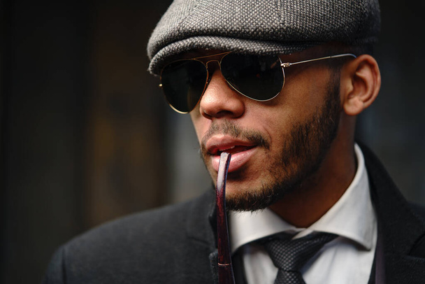 Studio shot of stylish african american man wearing coat, cap, glasses and holding smoking pipe - Photo, image