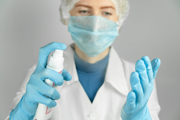 El médico con una bata blanca, máscara médica, gorra médica y guantes azules desinfecta sus manos con aerosol antiséptico de cerca - Foto, Imagen