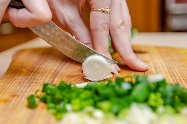 Γυναικεία χέρια κόβουν πράσινο κρεμμύδι με μαχαίρι σε ξύλινη σανίδα. υγιεινή διατροφή, μαγείρεμα, χορτοφαγικό φαγητό - Φωτογραφία, εικόνα