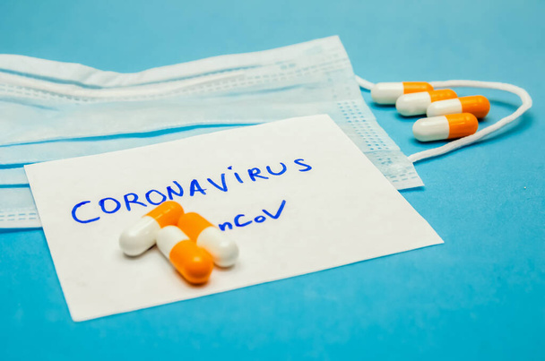 Medical mask, orange-white capsules on a blue background and the inscription Coronavirus on a white sheet close up. First aid, care during illness. - Foto, imagen
