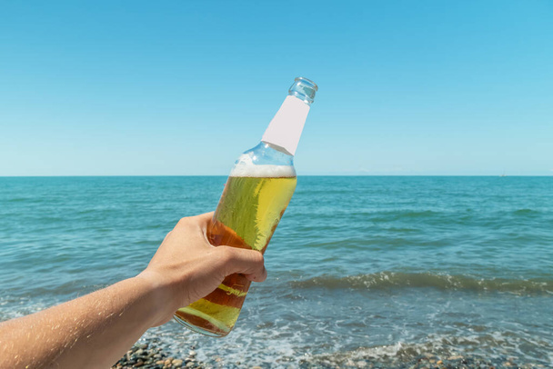 A man's hand holds a bottle of beer against the sea and blue sky on a Sunny day. The concept of rest. - Foto, imagen