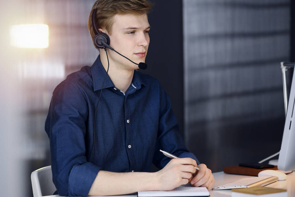 Young blond businessman using headset and computer in a darkened office, glare of light on the background. Startup business means working hard - Foto, immagini