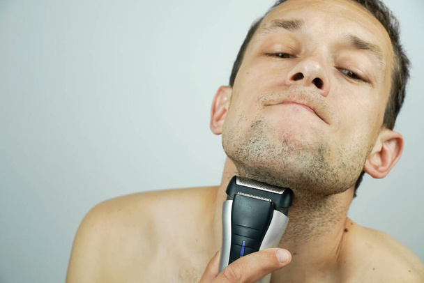 moreno hermoso hombre se afeita con una máquina de afeitar eléctrica y hace caras a la cámara. - Foto, Imagen