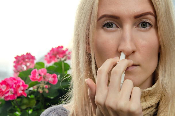 junge blonde Frau verwendet ein Nasenspray für eine laufende Nase oder Allergie vor einem Hintergrund von Zimmerblumen, Kopierraum. - Foto, Bild