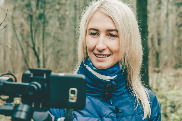 Молода блондинка-блогер знімає відео на мобільному телефоні і щупальце в лісі, посміхаючись і махаючи на камеру. концепція блогінгу, інформації, сучасних технологій
. - Фото, зображення