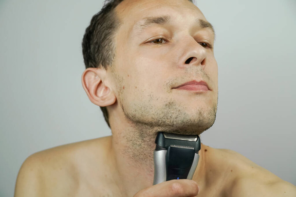 Porträt eines schönen dunkelhaarigen Mannes mit einem elektrischen Rasierer auf hellem Hintergrund. Dunkelhaariger Mann rasiert sich mit Rasiermesser in Großaufnahme. - Foto, Bild