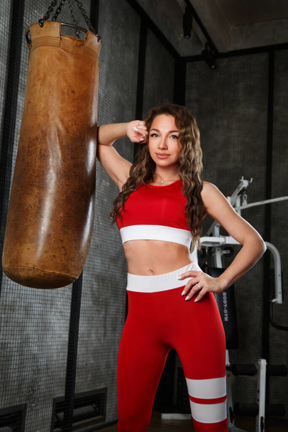 Nice girl in red sportswear in the gym near a punching bag. - Photo, image