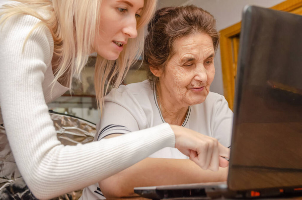 giovane ragazza bionda insegna a una donna anziana dai capelli scuri, la nonna come lavorare su un computer portatile a casa - Foto, immagini