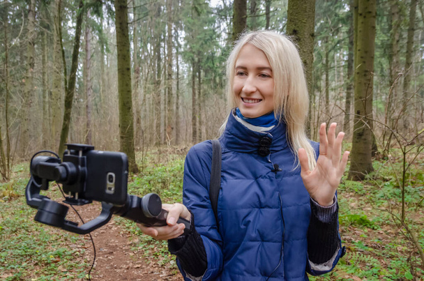 La joven bloguera rubia está filmando un video en un teléfono móvil y un gimbal en el bosque, sonriendo y saludando a la cámara. el concepto de blogging, información, tecnologías modernas. - Foto, Imagen