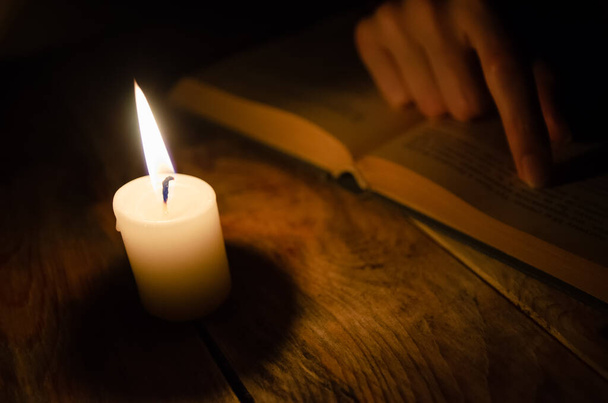 открытая книга, палец на книге, и горящая свеча на деревянном столе в темноте - Фото, изображение