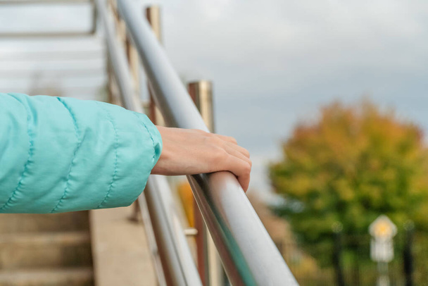 woman's hand in a turquoise jacket touches a metal railing against a background of yellow autumn trees close up - Фото, зображення