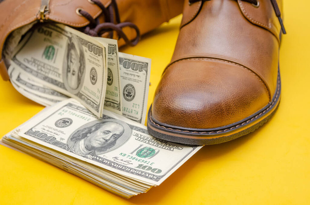 Concept photo de chaussures pour hommes en cuir marron, avec des dollars sur un fond jaune dans l'obscurité avec des lumières - Photo, image