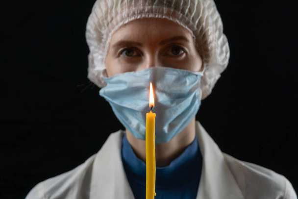 Νεαρή γυναίκα με ιατρική μάσκα, ιατρικό καπέλο και λευκό παλτό κρατά ένα αναμμένο κερί μπροστά της σε μαύρο φόντο. Έννοια της τελευταίας ελπίδας, πίστη σε ένα λαμπρό μέλλον. - Φωτογραφία, εικόνα