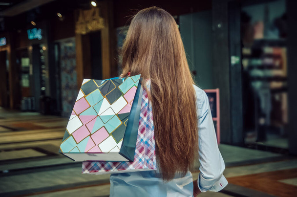 Красива молода дівчина з довгим волоссям у сорочці, тримає сумки, ходить навколо магазину, вигляд ззаду з рукою над плечима, порожнє місце на торговому мішку
 - Фото, зображення
