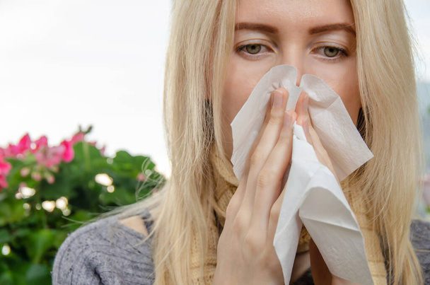 young blonde woman sneezes and covers her nose with a napkin against a background of indoor plants. Concept of health, medicine - Photo, Image